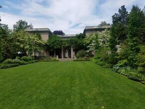A large manicured grassy lawn overlooks a traditional grey house. On both side of the cut grass are tall pine trees and small shrubbery outlining the entire yard. A variety of trees cover the surrounding yard. 