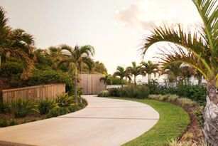 Picture of an edged driveway with green grass and groomed palm trees surrounding it. The sidewalk leads to the water, and on the surrounding sides of the sidewalk have a wooden fence. 
