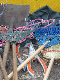 A picture of used garden tools consisting of a rake, large hedge clippers, garden gloves, and a shovel with dirt on it.
